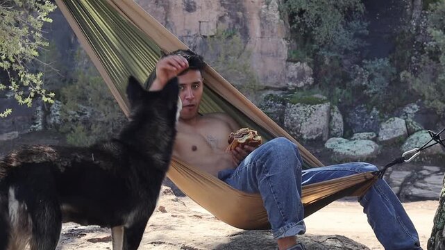 Shirtless man resting in hammock shares his meal with a black wolf-dog during a camping adventure in a rocky forest clearing, showcasing a beautiful bond between man and animal