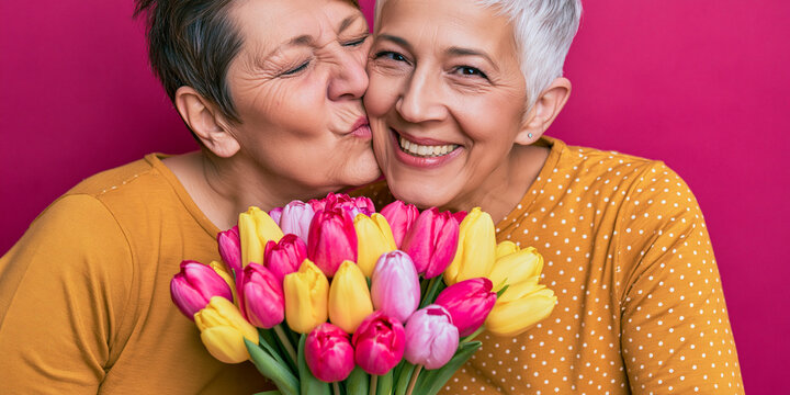 A heartwarming portrait of two women, one planting a gentle kiss on the other's cheek while holding a vibrant bouquet of tulips. Their expressions radiate happiness and affection.