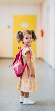 A young girl with pigtails and a bright yellow checkered dress carries a pink backpack, ready for school, with a warm smile