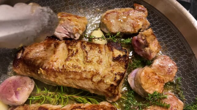 Sizzling veal steak and tender medallions fry in a hot skillet with fragrant herbs. A chef turns the meat using metal tongs, adding fresh thyme, rosemary, and whole garlic cloves.