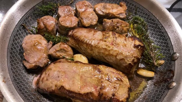 Cooked veal medallions sit seared in a hot pan after frying, showcasing thick tenderloin steaks infused with fresh rosemary, whole garlic cloves, and aromatic thyme.