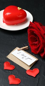 Woman putting dessert in shape of heart, card with phrase I Love You, red hearts and rose at black table, closeup