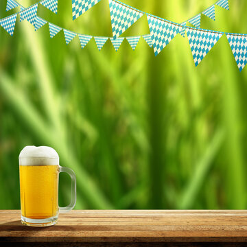 A glass of cold beer with thick foam on a rustic wooden table against a blurred green background, decorated with Bavarian blue and white checkered flags for Oktoberfest. Bavarian Oktoberfest Festival