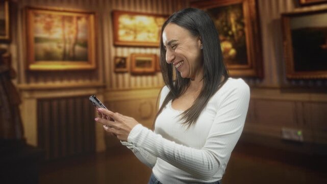 Woman smiling and texting on smartphone, hands visible, wearing white top with framed paintings behind in a gallery building; joy connection.