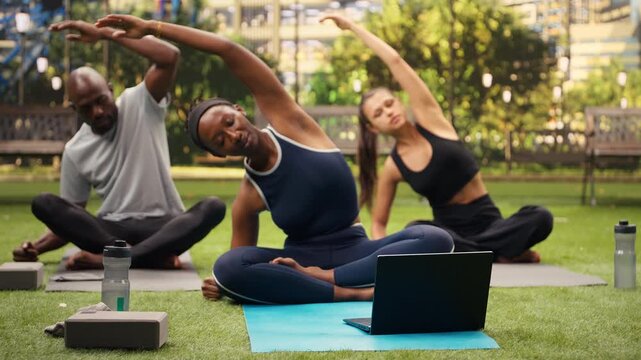 Young woman does side stretch exercise in front of laptop with friends, following a digital yoga activity session with participants in city park while stretching together on mats for flexibility.
