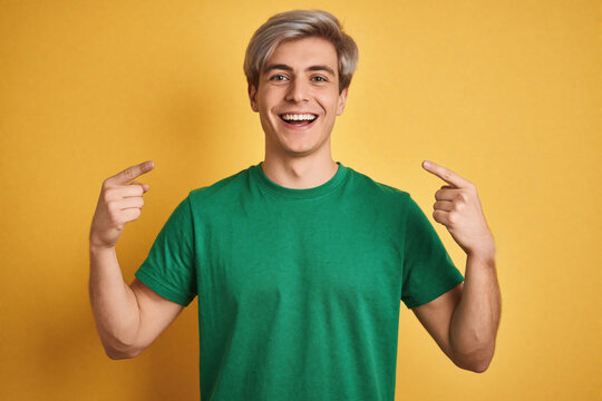 Handsome young man in green t-shirt smiles, points at. Cheerful caucasian male with blonde hair poses against bright yellow background. Confident person shows pride, success with happy expression.