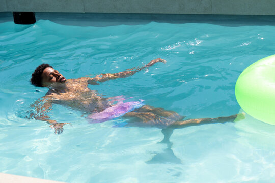 African American man floating on back in backyard pool in purple-pink trunks with lime-green ring