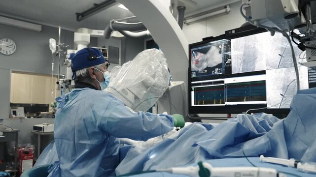 Interventional doctor working beside a patient, C-arm system and angiography monitor during a catheter procedure.