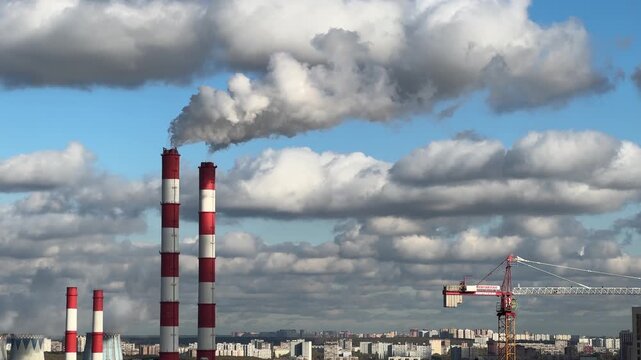 Red and white industrial chimneys releasing white steam into a blue sky with fluffy clouds and a construction crane in Moscow in Russia