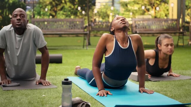 Yoga instructor and participants hold cobra pose on mats in city park, practicing flexibility together during an outdoor backbend Pilates exercise.