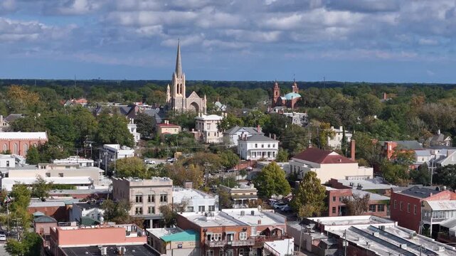 Town view with church steeple rising above rooftops. Blue sky with clouds above green trees. Buildings mix brick and white paint. Peaceful quiet scene, no people visible. Good for tourism, real estate