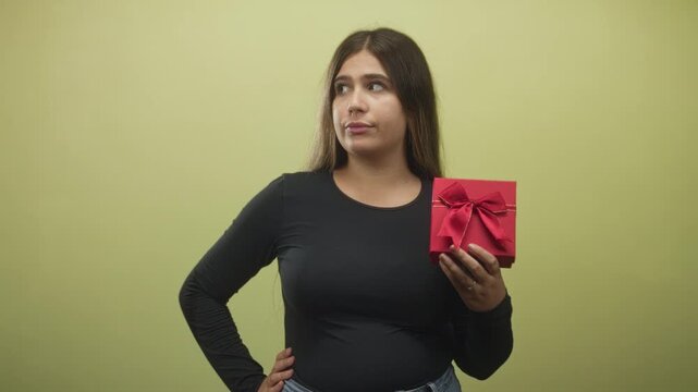Woman holding red gift box with hand on hip and pouting expression against plain green studio wall; mild annoyance.
