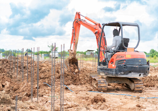 Small orange excavator digging soil at an outdoor construction site with foundation rebar steel rods in the foreground.
