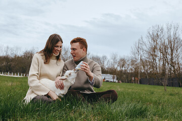 Obraz premium A couple sits on green park grass in cozy sweaters, sharing a playful moment with a fluffy dog, enjoying a relaxed outdoor afternoon together in natural light