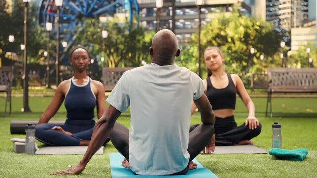 Yoga instructor demonstrates a seated side neck stretch as participants follow on mats in city park, supporting mobility and guided wellness exercise.