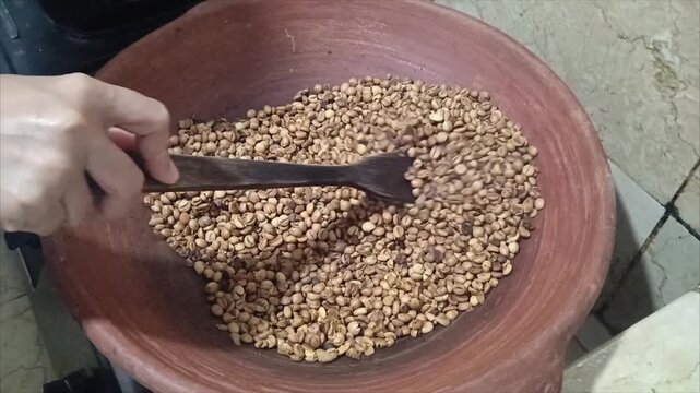 Top view of hand stirring and roasting coffee beans in traditional clay earthenware pan on stove, traditional manual coffee roasting process in Indonesia
