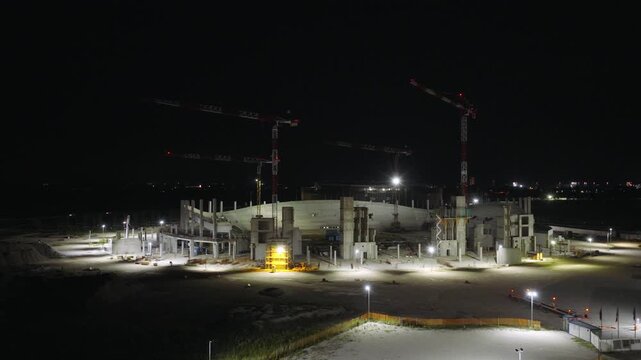 Concrete support columns and curved stands rise under tall red cranes at new Venezia FC stadium site, floodlit construction zones glow against dark Venice night sky. Aerial drone approaching shot