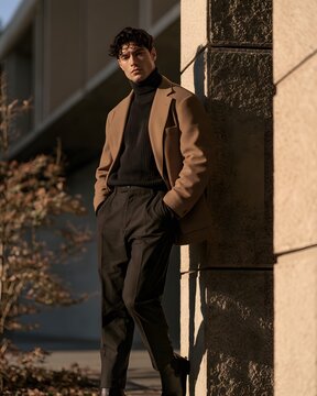 Stylish man in a beige blazer and black turtleneck leaning against a stone wall during golden hour