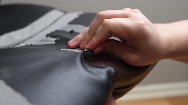 A person peels a cracked, flaking synthetic leather layer from a black chair seat indoors, exposing the grey backing underneath. A concept of furniture restoration and worn material renewal.