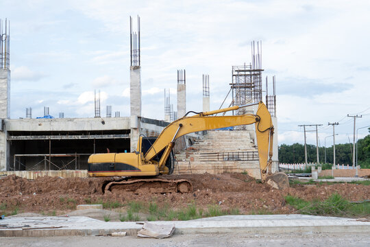 Yellow excavator digger performs earthwork on dirt ground near concrete pillar at building construction site, showing powerful machinery drive with ambitious focused energy for land development