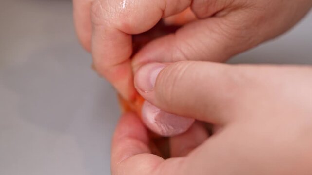 Human hands carefully pull the thin orange wrapper off a pink cooked sausage, gradually uncovering the smooth meat on a gray countertop, a concept of food preparation and snack time.