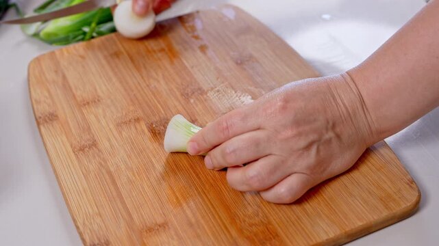 A woman hand holds a spring onion bulb steady and cuts away its green stalk with a knife on a bamboo board, a simple kitchen skill for batch prepping farm market produce at home.