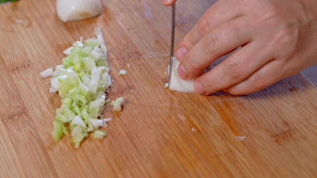 A hand holds a peeled onion steady and works a blade through it until only a neat mound of tiny pieces remains next to sliced green stalks, a lesson in home knife skills.