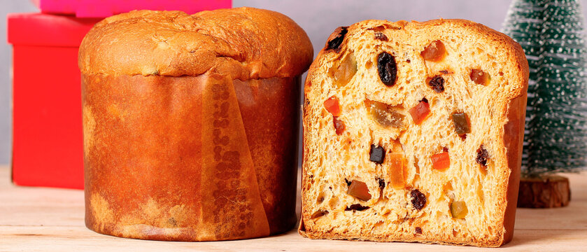 Whole and sliced Christmas panettone with candied fruit and raisins on a wooden surface with gift boxes and festive decorations.
