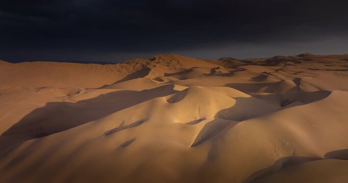Vast desert sand dunes illuminated by sunlight under dark moody sky in Huacachina, Peru. Aerial forward