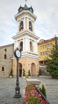 Vertical view of an antique double-faced post clock in a Plovdiv square with the Holy Mother of God Church bell tower behind.