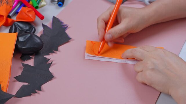 Hands hold an orange felt tip pen and sketch a design on layered orange and white napkin paper, beside black paper bat cutouts and balloons on a pink desk, a concept of festive DIY decor.