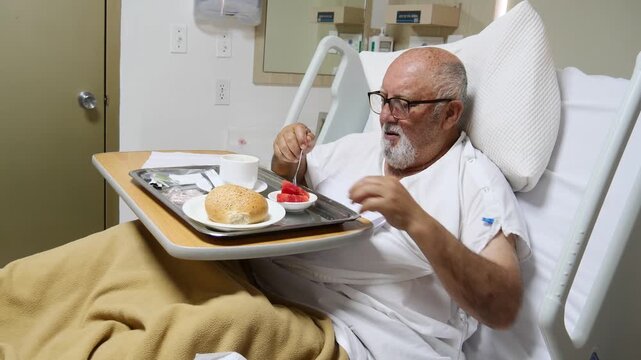 Elderly man wearing a hospital gown eats breakfast while resting in a hospital bed. A meal tray with bread, tomato, a cup, utensils, and napkins is positioned on an adjustable bedside table in the pat