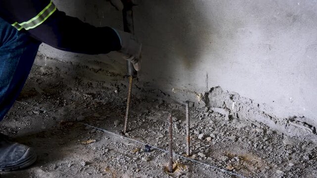 Construction worker using a heavy hammer to drive metal rods into the ground at a building site. The activity shows manual groundwork and preparation for an electrical installation or grounding system