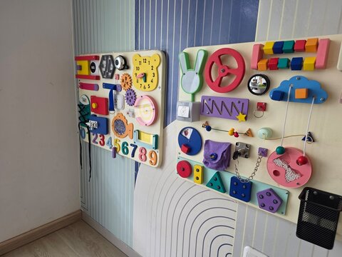 A static shot of two colorful educational sensory boards, or busy boards, mounted on a white and blue patterned wall inside a children's room or playroom.