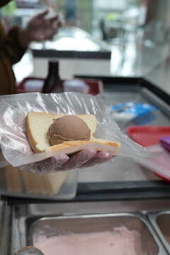 Chocolate ice cream scoop served on white bread slice held by gloved hand with clear plastic wrap in street food stall for dessert snack.