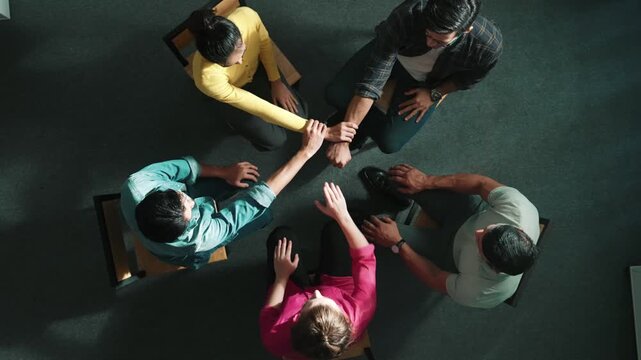 Top down aerial view of diverse business people hold wrist while sitting circle together. Faithful prayers holding wrist or hand of each other. Represented teamwork, unity, togetherness. Symposium.