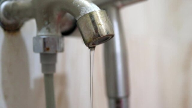 A weak stream of water dripping slowly from a household metal faucet inside a bathroom during a crisis caused by AI data center consumption