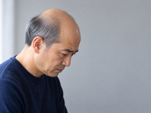 グレー背景で斜め横向きにうつむく紺色のセーターを着た白髪混じりの50代日本人男性と薄い頭頂部