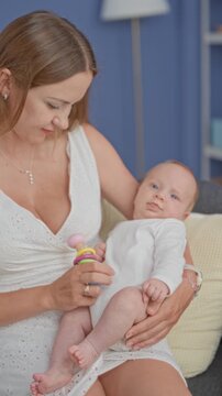 Mother holding newborn baby with bare feet and colorful rattle in home setting; tenderness bonding.