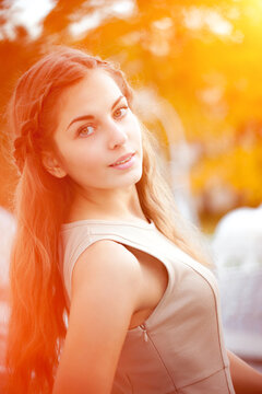 Woman with long reddish-brown hair and a braided side section, wearing a sleeveless beige dress, looking over her shoulder with a soft smile against a blurred orange background.