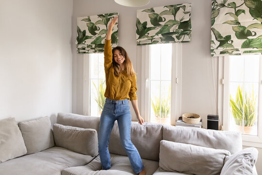 Adult Hispanic woman listening favourite music, dancing with raised arms on sofa in modern living room, having fun at new house, rented apartment or own flat, enjoying carefree leisure alone indoors
