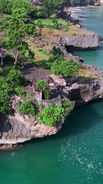 Aerial view bali temple cliff overlooking turquoise ocean, drone captures cliffside shrine surrounded by lush greenery, rocky cove and emerald lagoon, waves lapping against stone, vertical video