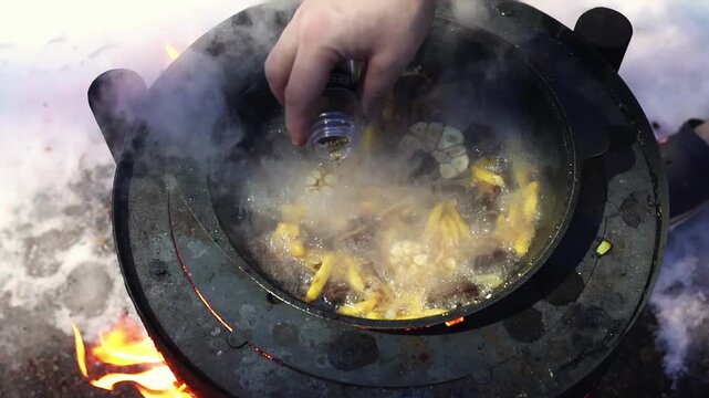 Traditional plov simmering outdoors in winter while a person seasons the dish by hand. Rice, meat, garlic and carrots cook over a wood burning stove in snowy weather. Pilaf boiling.