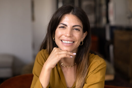 Closeup of smiling adult Hispanic woman relaxing alone in cozy apartment, looking at camera, posing for portrait, showing positivity, confidence, enjoying comfort and weekend rest indoors. Wellbeing