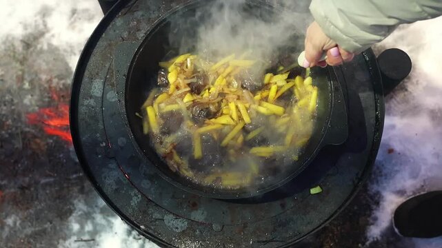 Close-up of plov simmering over a fire in snowy weather, with rice, broth and spices being stirred by hand. Traditional outdoor cooking and winter meal preparation concept.
