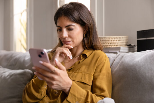 Focused Hispanic woman sitting on sofa checking smartphone, chatting online, reviewing information, browsing social media, scrolling through platforms, reading message, news, articles or blogs at home