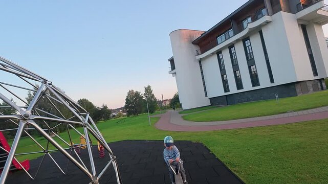 Young boy enjoys a playful moment on a seesaw in an urban playground, surrounded by modern buildings and lush green grass in the warm sunset light, youthful joy and energy.