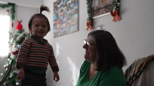 A laughing toddler points at something while standing beside her smiling grandmother near a decorated Christmas tree at home