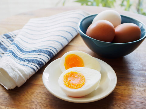 Soft boiled egg cut in half on a white plate with blue striped linen cloth and eggs in a bowl on a wooden table by the window