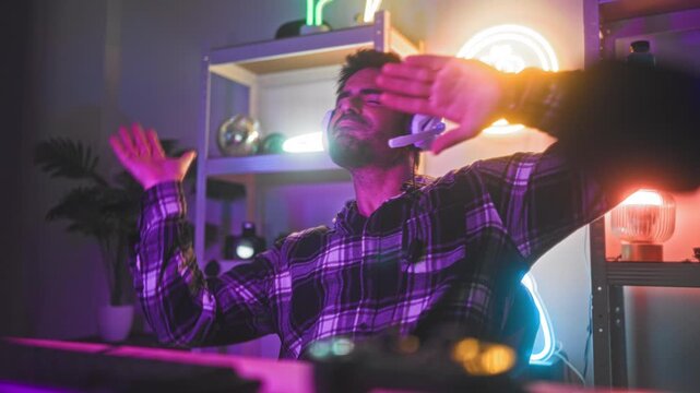Man in headset and plaid shirt dancing with hands raised and eyes closed beside neon bitcoin sign in a streaming studio; excitement celebration.